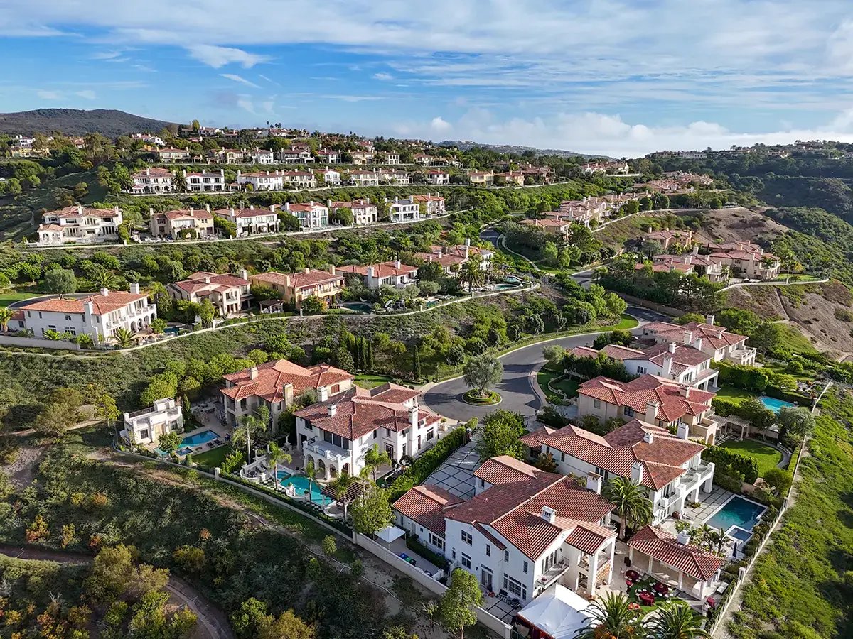 Aerial view of a luxurious hillside neighborhood with Mediterranean-style homes featuring terracotta roofs, lush greenery, winding roads, and blue skies