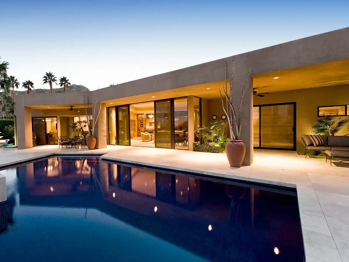 Modern house exterior with large windows opening to a poolside patio at dusk